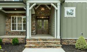 Front entrance of a house with stone and green siding, a covered porch supported by white columns, a double wooden door with glass panels, and steps leading up to the porch. Small shrubs are planted on either side.