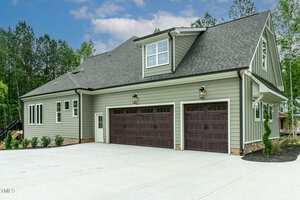 A modern two-story house with green siding and a large driveway. The home features three wooden garage doors, multiple windows, and is surrounded by trees and landscaped greenery.