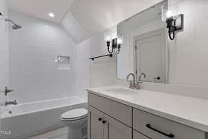 A modern bathroom with white cabinetry, a large mirror, two wall-mounted light fixtures, a white countertop, a toilet, and a bathtub with white subway tile walls and a built-in niche.