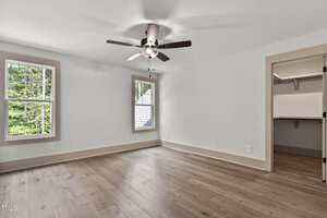 A bright, empty room with light-colored walls, two windows, wood flooring, a ceiling fan with light, and an open doorway leading to a walk-in closet with built-in shelving.