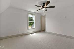 Empty room with light gray carpet, white walls, a single window showing greenery outside, a ceiling fan with lights, and an open doorway leading to another space. Natural light enters through the window.