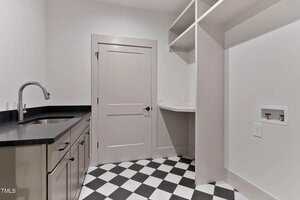 A laundry room with black and white checkered tile floor, light-colored cabinets with a black countertop and sink, a built-in desk, open shelves, and a door in the center of the back wall.