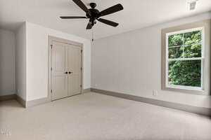 Empty bedroom with beige carpet, light-colored walls, double-door closet, ceiling fan with dark blades, and a window showing green trees outside.