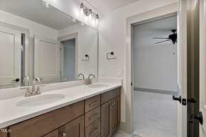 A modern bathroom with a double vanity, two sinks, large mirror, light fixtures, and wood cabinets. An open door leads to a carpeted room with a ceiling fan. The walls and countertops are light-colored.