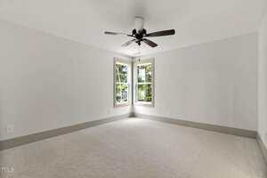 Empty room with light-colored carpet, white walls, a ceiling fan with lights, and two windows in the corner letting in natural light. The room appears clean and well-lit.