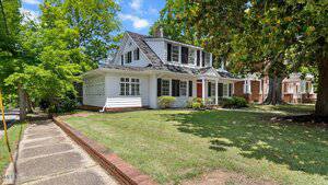 A white two-story house at 830 S Chestnut Street in Henderson features a red front door, large windows, and a covered porch on a corner lot with green grass, shrubs, and mature trees under a blue sky with scattered clouds.