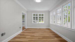 A bright, empty room with light gray walls, white trim, and wood-look flooring. Large windows line two walls, letting in natural light and providing views of greenery outside. A door leads to another part of the house.