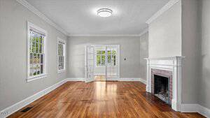A bright, empty living room with hardwood floors, white walls, multiple windows, a white fireplace, and glass French doors leading to another sunlit room. A round ceiling light is centered above.