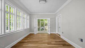 Empty room with light gray walls, white trim, multiple tall windows on the left wall, and double glass doors leading to an outdoor area. The floor is light wood, and the ceiling has a modern circular light fixture.