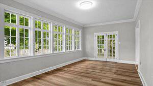 A bright, empty room with light gray walls, wood flooring, large windows on one side, and white French doors on the opposite wall. Natural light fills the space, and green trees are visible outside.