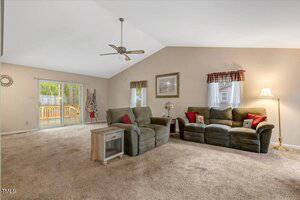 A living room with beige carpet, olive green couch and loveseat, small side table, floor lamp, wall art, and ceiling fan. Two windows with valances and sliding glass doors open to a wooden deck outside.