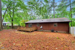 A single-story brown house with a sloped roof and a wooden deck in a fenced backyard surrounded by trees and pine needles covering the ground.