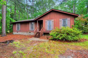 A one-story brown house with wood siding at 506 E E Street in Butner, featuring three front windows, a small porch with steps, and surrounded by trees and shrubs on a pine needle-covered yard.