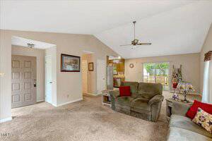 Living room with beige walls and carpet, green upholstered couch and loveseat, red accent pillows, ceiling fan, large window, kitchen area with wooden cabinets, and sliding glass door leading to a deck.