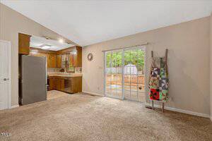 A carpeted living area with beige walls opens into a kitchen with wood cabinets and stainless steel appliances. Sliding glass doors lead to a fenced backyard. A ladder with quilts leans against the wall.