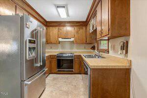 A small kitchen with wood cabinets, a stainless steel refrigerator, an electric stove with a range hood, a double sink by a window, laminate countertops, and beige tile flooring.