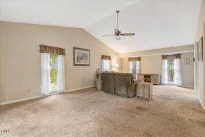A carpeted living room with beige walls, vaulted ceiling, ceiling fan, sofa, side table, desk, and three windows with valances. A framed picture hangs on one wall. The room is well-lit with natural light.
