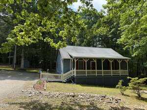 A blue house with a covered front porch and white railings sits in a wooded area. The yard has grass, trees, and a gravel driveway. The sky is partly cloudy.