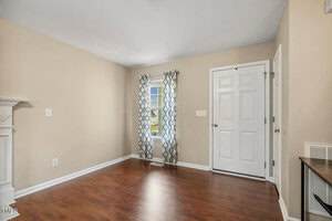 A small, empty room with beige walls, hardwood floors, a white-paneled front door, a window with patterned curtains, and a white trim along the baseboards and door frames.