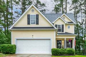 A two-story yellow house with black shutters, a double garage, and well-maintained landscaping on 1984 Sadler Avenue in Creedmoor. Tall pine trees and overcast skies frame the home, with a hanging basket of purple flowers by the entrance.