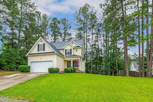 A two-story yellow house with white trim and a gray roof sits on a large, green lawn. The home has a double garage and is surrounded by tall pine trees on a partly cloudy day.