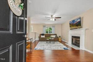 A living room with wood flooring, a brown sectional sofa, two round coffee tables on a patterned area rug, a fireplace with a TV above it, beige walls, and a ceiling fan. The door is open with a "Welcome" sign.