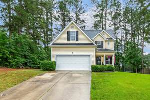 A two-story beige house with white trim and a double garage sits at the end of a concrete driveway, surrounded by green grass, shrubs, and tall pine trees under a partly cloudy sky.