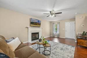 A living room with beige walls, wood floors, a ceiling fan, a gray sectional sofa, a fireplace with a TV above it, two round coffee tables, a patterned rug, and a door with sidelights.