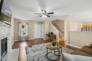 A living room with a gray and white area rug, round coffee tables, a brown sofa, and a ceiling fan. There is a fireplace with a TV above it, a staircase, and a view of the kitchen in the background.