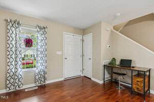 A small room with light brown walls and wood flooring features a window with patterned curtains, a white front door, a small desk with a laptop and chair, and a staircase leading upstairs.