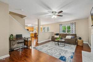 A living room with wood flooring, a sectional sofa, blue and white rug, and a round coffee table. A staircase, home office desk, and kitchen with dining area are visible in the background. Neutral-colored walls and ceiling fan.