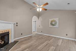 A room with light wood flooring, beige walls, a white ceiling fan with lights, a fireplace on the left, an arched doorway, and a decorative wall art piece on the right wall.