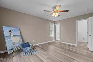 A clean, empty room with tan walls, wood floor, ceiling fan with lights, two windows, two open white doors, and a painting of a woman in a white dress on an easel beside a blue blanket and pillow.