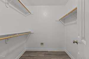 Empty walk-in closet with white walls, wooden rods, and shelves on both sides. The floor is wood-look laminate, and there is a small square wall vent near the floor at the back. The door is partially open.