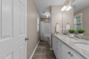 A modern bathroom with light brown walls, a double-sink marble countertop, white cabinets, wood floors, two mirrors, two small potted plants, towel rings, and a shower area with a white curtain in the back.