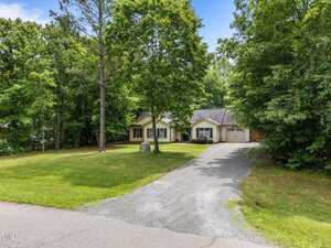 Single-story house with light-colored siding and dark shutters sits surrounded by large trees and greenery. A gravel driveway leads up to the attached garage. The lawn is spacious with a stone marker near the front.