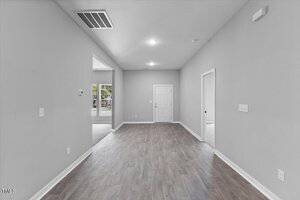 A bright, empty room with gray walls, white trim, and wood-style flooring. An open doorway leads to another room on the left, with a closed white door at the far end and a smaller open door on the right.