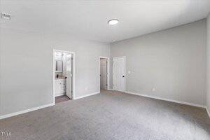 A spacious, empty room with gray carpet, light gray walls, a round ceiling light, and two doors leading to a bathroom and a closet. The bathroom features a vanity with a mirror.