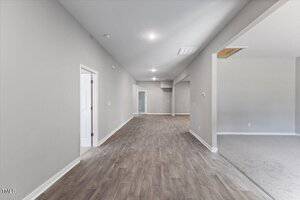 A hallway with wood-look flooring leads to a distant doorway; gray walls and ceiling lights are visible, with an adjacent carpeted room on the right separated by a partial wall.