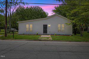 Charming single-story gray house at 714 Harriett Street in Henderson, featuring a black front door and two large windows, set against a lawn with trees at sunset. A mailbox sits to the left near the street, with a paved walkway leading to the entrance.