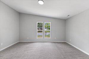 A vacant room with light gray walls, beige carpet, a ceiling light, and two rectangular windows letting in natural light and showing a view of the street and greenery outside.