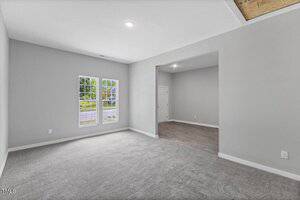 A room with gray carpet, light gray walls, and a large window facing outside. The room leads into another area with wood flooring and a white front door visible in the background. Ceiling light is turned on.