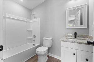 A modern bathroom with a white bathtub and shower, a toilet, and a white vanity with a granite countertop beneath a mirror. The space has light-colored walls and wood-style flooring.