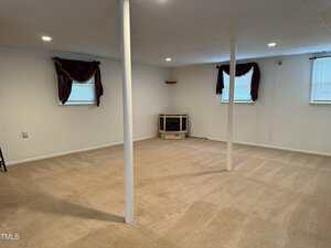 A mostly empty, carpeted basement room with white walls, two support poles, two small windows with dark curtains, a wall-mounted heater in the corner, and ceiling lights.