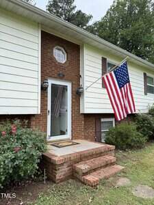 A two-story house with white siding and red brick exterior. A large American flag hangs by the front door, which has glass panels. There are shrubs, flowers, and steps leading to the entrance.