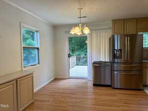 A kitchen with light wood flooring, beige cabinets, a stainless steel refrigerator and dishwasher, a white countertop, and a sliding glass door leading to a wooden deck outside. A light fixture hangs from the ceiling.