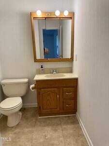 A small bathroom with a toilet on the left, a wooden vanity with a sink and mirror above it, and three lights above the mirror. The walls are white and the floor has beige tiles.