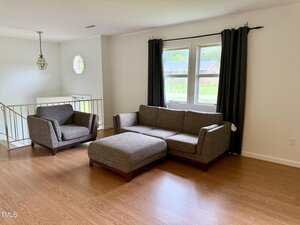 A living room with wood flooring, a gray sofa, matching armchair, and ottoman. A window with black curtains lets in natural light. A small chandelier hangs near a railing by a split-level entry.
