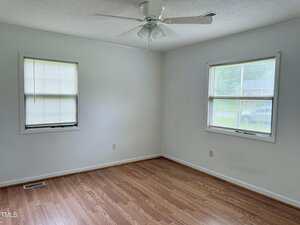 Empty room with light-colored walls, two windows with blinds, a white ceiling fan, and wood laminate flooring. The room has baseboards and electrical outlets, and natural light is coming in through the windows.