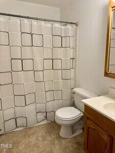 A bathroom with a white shower curtain featuring a black square pattern, a white toilet, a wooden vanity with a sink, a mirror above the sink, and light-colored flooring.